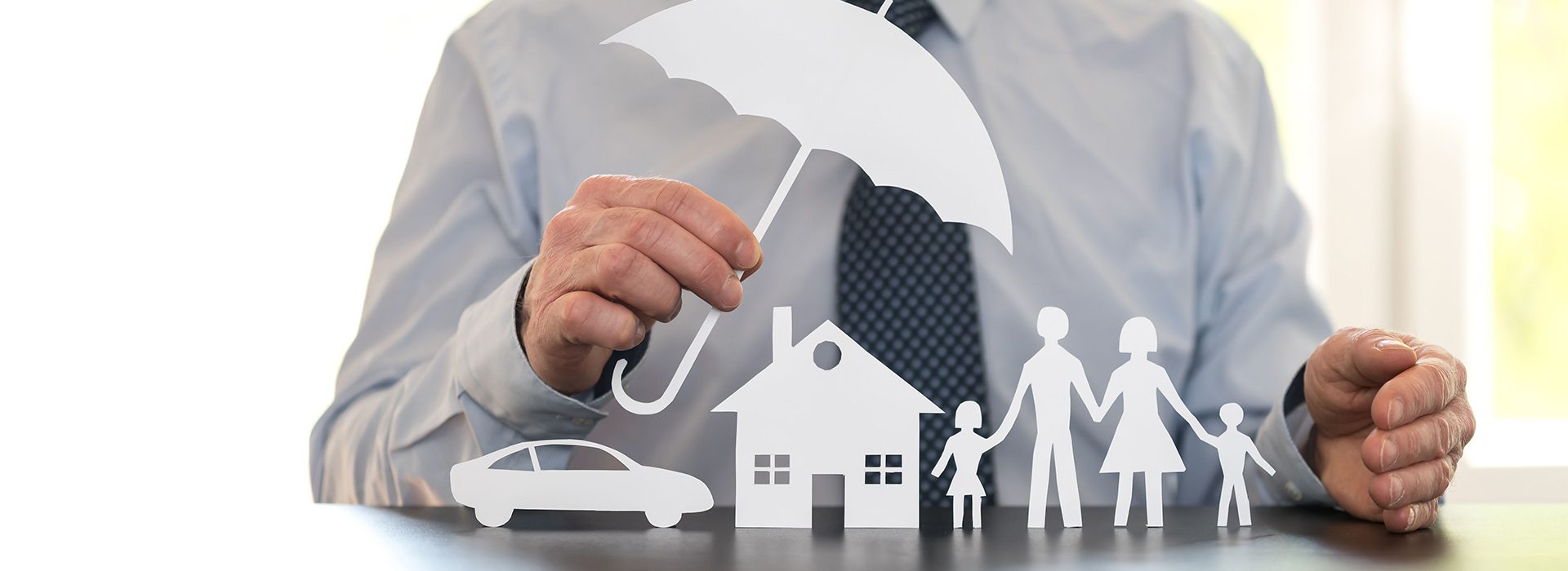 Personal-Umbrella-Insurance-Man-Holding-a-Paper-Umbrella-Over-Paper-Cutouts-of-a-Home-Car-and-Family-While-He-Sits-at-a-Desk